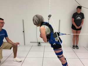 A woman in a harness leans forward toward a man. A student observes in the background.