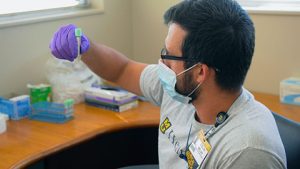 A white man wearing a mask and purple rubber gloves examines a vial of blood