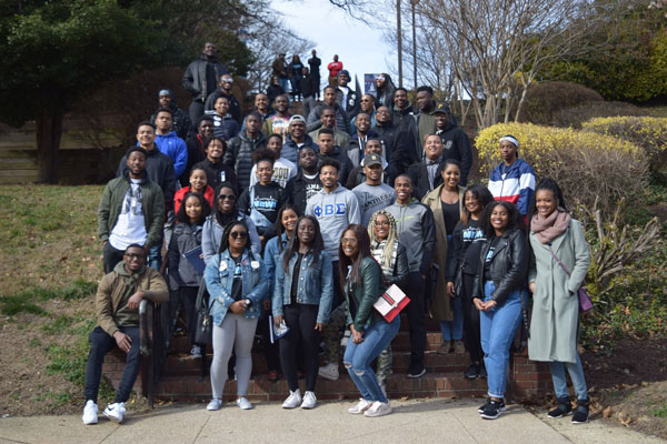 A large group of Black men and women in casual attire pose for the camera on a large outdoor concrete stairway