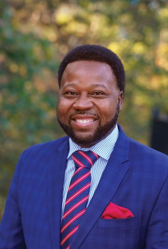 Portrait of a Black man wearing a blue blazer with blue-and-red striped tie and red pocket handkerchief.